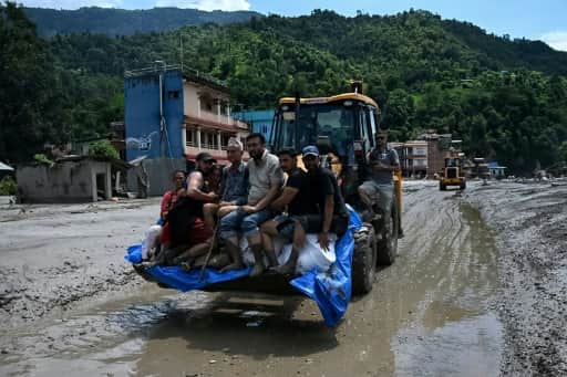 Des habitants évacués assis dans le godet d'un engin de chantier au milieu d'une rue couverte de boue