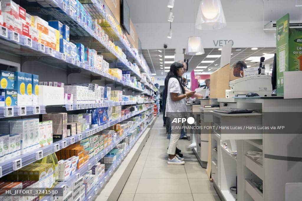 Des rayonnages de médicaments dans une pharmacie, une cliente au comptoir