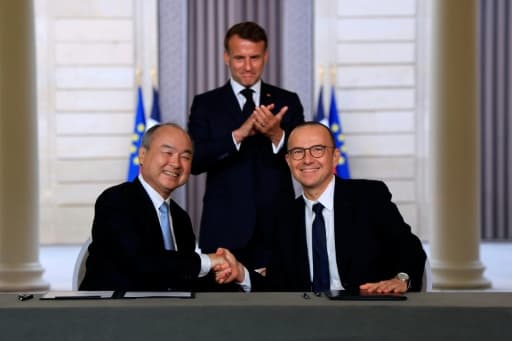 Deux hommes se serrent la main devant des documents, un troisième applaudit debout derrière eux