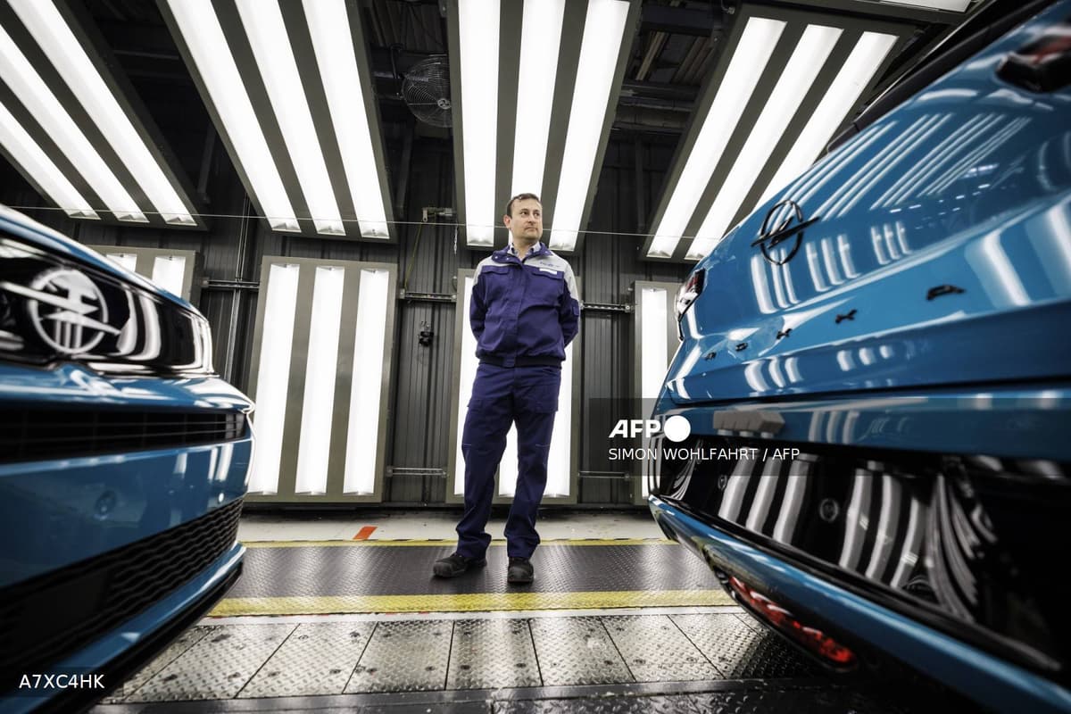 Un ouvrier Stellantis en uniforme bleu debout entre deux Opel bleues sur la chaîne d'assemblage de l'usine de Poissy, sous les lumières néon caractéristiques des sites de production automobile