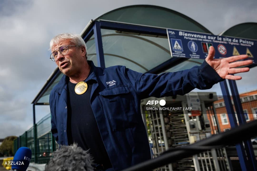 Un syndicaliste en uniforme PSA Groupe, portant un badge « NON À LA FERMETURE DE STELLANTIS POISSY », s'exprime devant l'entrée du site, sous le panneau « Bienvenue sur le site de Poissy »