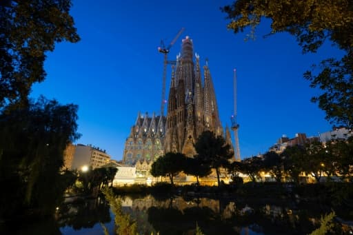 La basilique de la Sagrada Família d'Antoni Gaudí à Barcelone, illuminée au crépuscule, avec ses grues de chantier et son reflet dans un bassin