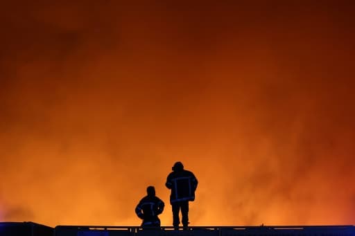 Deux sapeurs-pompiers en silhouette face aux fumées orange de l'incendie
