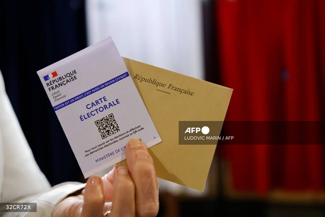 Une main tient une carte électorale de la République française et une enveloppe de vote devant un isoloir tricolore