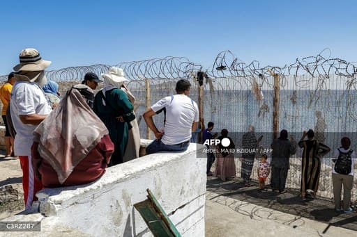 Des personnes debout derrière une clôture surmontée de barbelés qui descend jusqu'à la mer