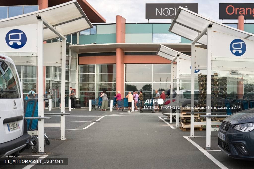 Des clients poussent des chariots sur le parking d'un centre commercial, devant l'entrée du magasin
