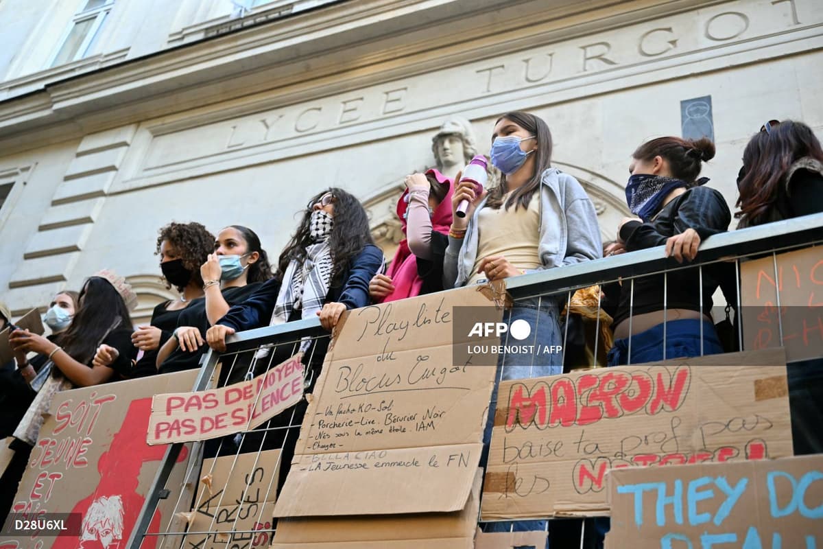 Des jeunes au visage en partie couvert se tiennent derrière une barrière garnie de pancartes en carton, sous une façade où est gravé « Lycée Turgot »