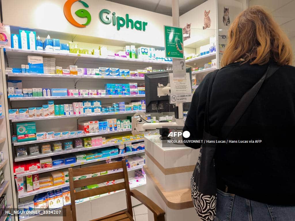 Une femme de dos au comptoir d'une pharmacie, devant des rayons de médicaments et de produits de soin