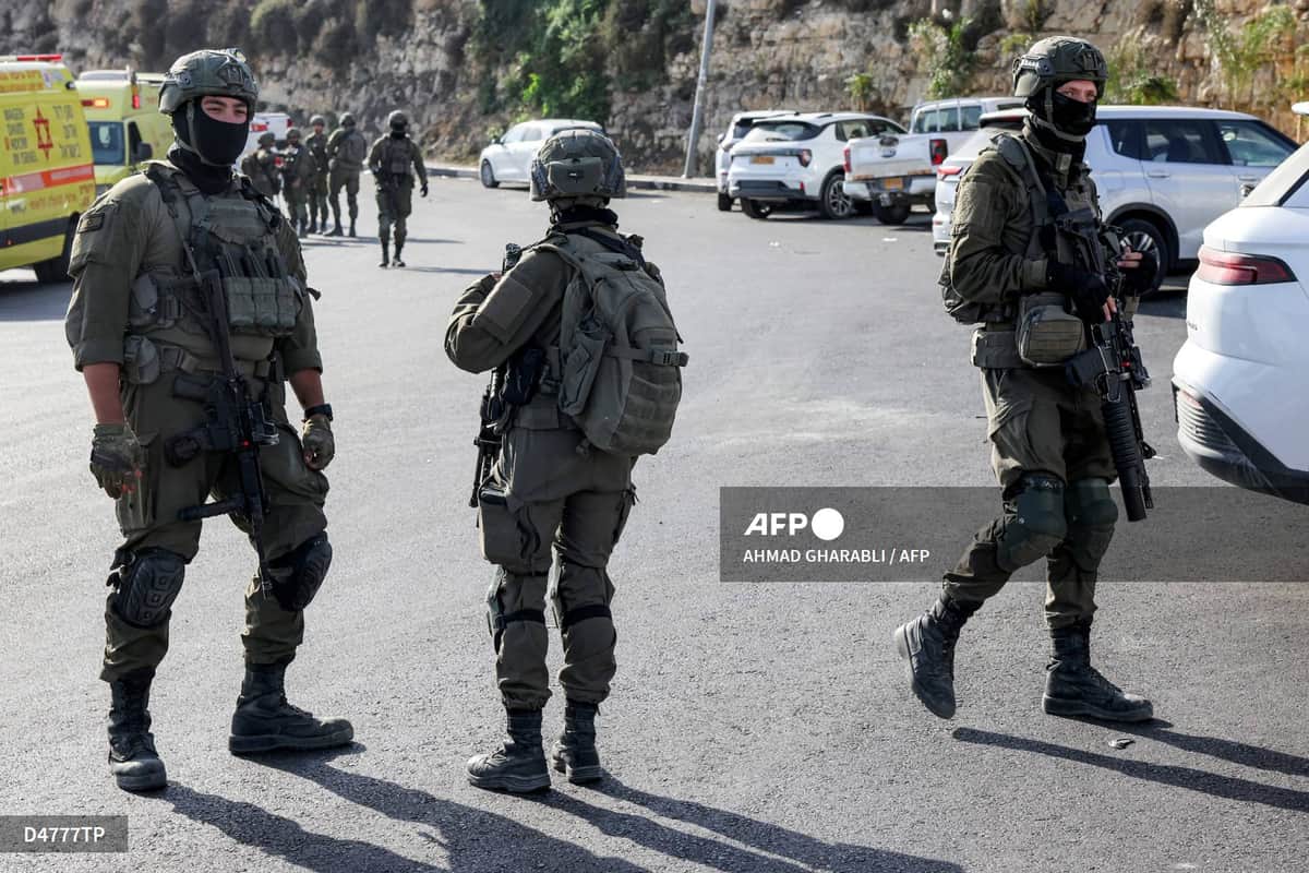 Trois soldats israéliens casqués et armés sur une route, des ambulances jaunes en arrière-plan
