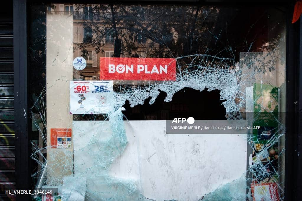La vitrine d'un magasin brisée, percée d'un large trou, sous une affiche « Bon plan »